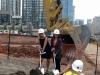 Two women digging into sand with contruction excavator in background.