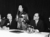 Jean Lumb speaking with microphone at conference table with four men
