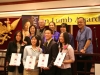 group posing for photo together on stage, front row of students are holding their award plaques