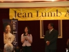 women speaking into microphone next to Jean Lumb banner