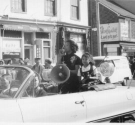 Jean Lumb sitting in back of converitble down busy pedestrian street in chinatown holding a megaphone. Young girl beside her holding a lantern.