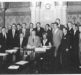 Jean Lumb sitting at long conference table signing documents. large group of male chinese delegates grouped behind table